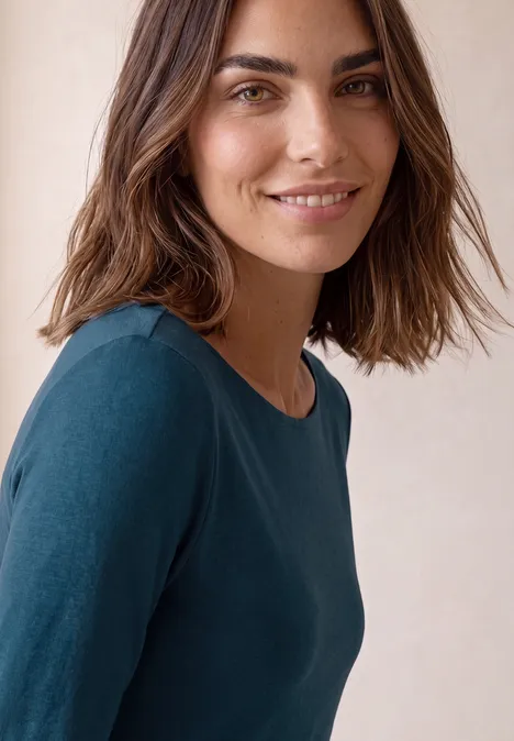 Porträt einer lächelnden Frau mit welligen brünetten Haaren und blauem Oberteil vor hellem Hintergrund.