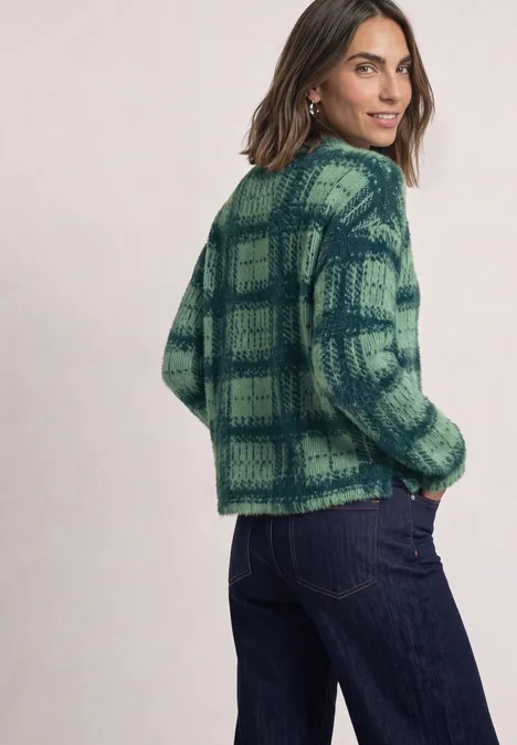 Eine Frau trägt eine grün-blaue karierte Strickjacke und Jeans, sie schaut über die Schulter.