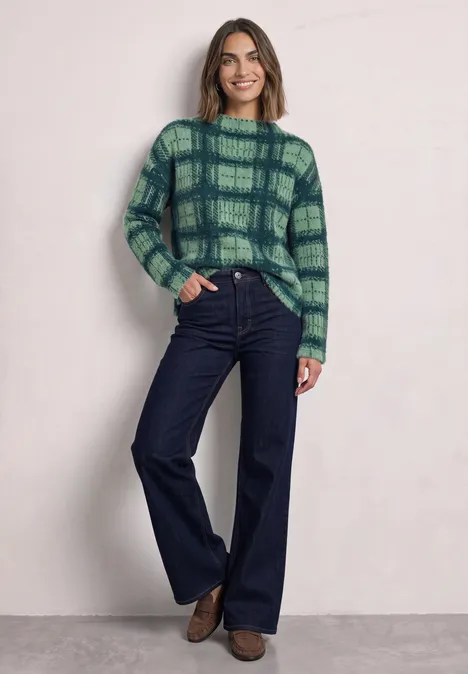 Eine Frau trägt einen grünen karierten Pullover und weite Jeans vor hellem Hintergrund.