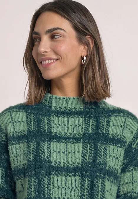 Frau mit braunen Haaren, trägt karierten grünen Strickpullover und silberne Ohrringe, lächelnd.