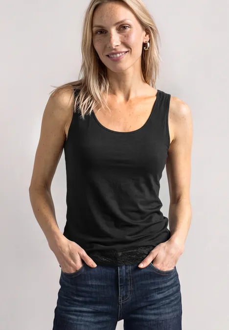 Lächelnde Frau trägt schwarzes Top und Jeans, steht vor hellem Hintergrund mit Händen in den Taschen.