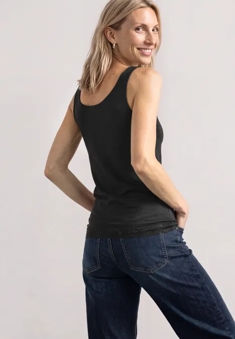 Frau in schwarzem Top und blauen Jeans, lächelnd, leicht zur Kamera gedreht, Hände in den Gesäßtaschen.