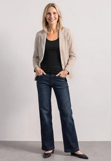 Eine lächelnde Frau steht in blauer Jeans, schwarzem Oberteil, beigem Blazer und schwarzen Schuhen.