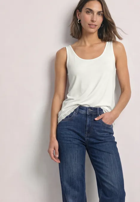 Eine Frau steht lässig mit einer Hand in der Jeanstasche, trägt weißes Tanktop und blaue Jeans.