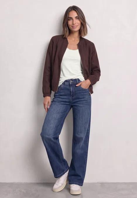 Frau in brauner Jacke, weißem T-Shirt, weiten Jeans und weißen Sneakern vor hellem Hintergrund.