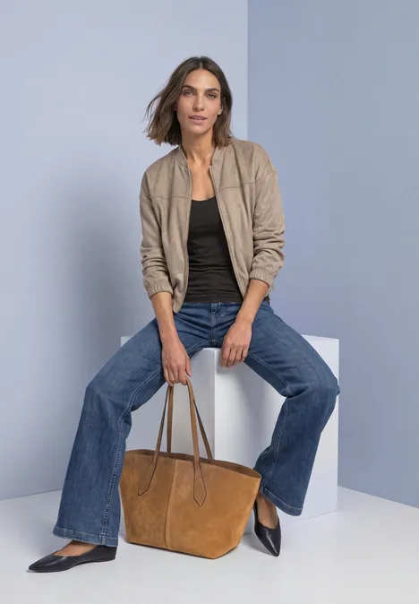 Frau sitzt auf weißem Würfel, trägt beige Jacke, schwarzen Top, weite Jeans und hält braune Tasche.