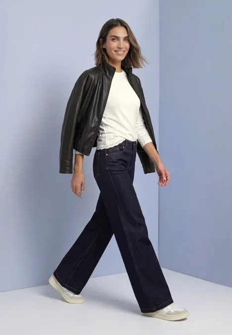 Frau mit weitem Jeans, weißem Shirt und Lederjacke über der Schulter, die selbstbewusst lächelnd geht.
