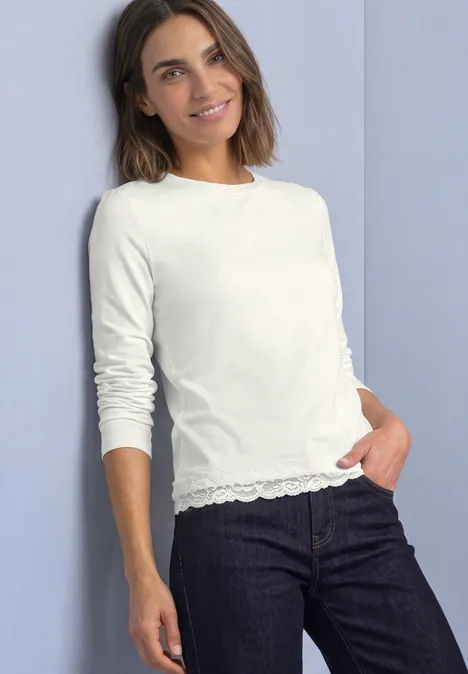Frau in schlichter weißer Langarmshirt mit Spitzenabschluss und dunklen Jeans vor hellem Hintergrund.
