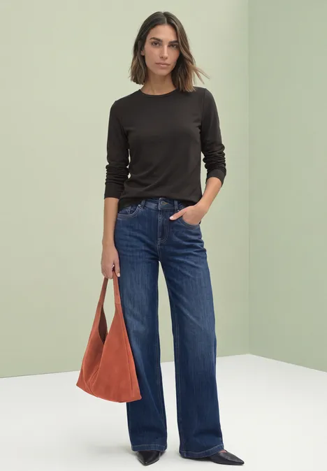 Modell in schwarzem Langarmshirt, weiten blauen Jeans und orangefarbener Tasche vor neutralem Hintergrund.