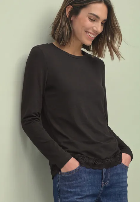 Frau in schwarzem Langarmshirt und blauen Jeans lehnt entspannt mit Händen in den Taschen.