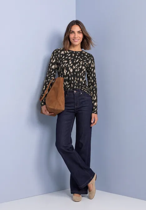Langarm Shirt mit Rundhals und Rehmuster Black Frau in gemusterter Bluse, dunkelblauen Schlagjeans und beiger Tasche vor pastellblauem Hintergrund.