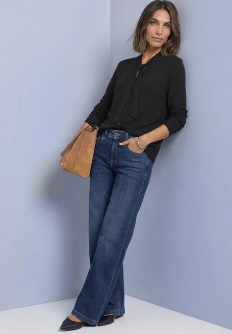 Langarm Shirt im Blusen-Look Black Frau lehnt an Wand; trägt schwarze Bluse, weite Jeans und halbrunde braune Tasche.