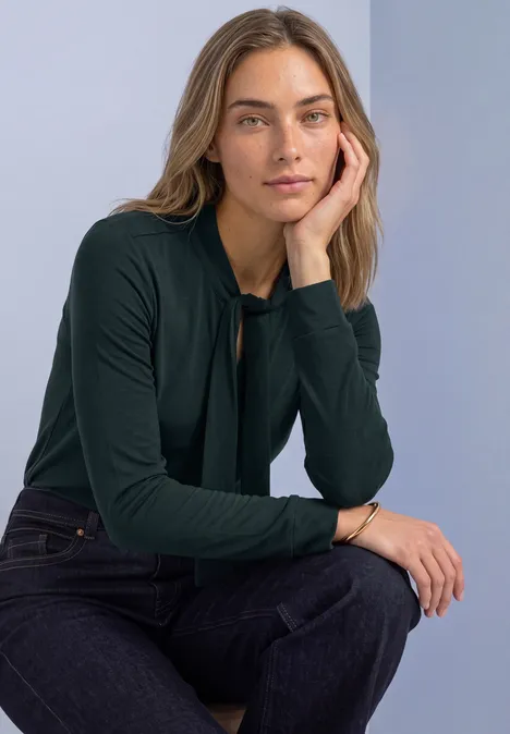 Shirt met lange mouwen in blouse-stijl stormy green Vrouw in donkergroene top en spijkerbroek leunt op haar arm en kijkt ontspannen in de camera.