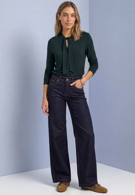 Shirt met lange mouwen in blouse-stijl stormy green Vrouw in groene cardigan, wijde donkerblauwe jeans en bruine schoenen voor een effen tweekleurige achtergrond.