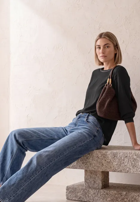 2-in-1 Sirt aus Chiffon mit Boat Neck Black Junge Frau sitzt lässig auf einer Steinbank, trägt schwarzen Pullover, Jeans und eine braune Schultertasche.