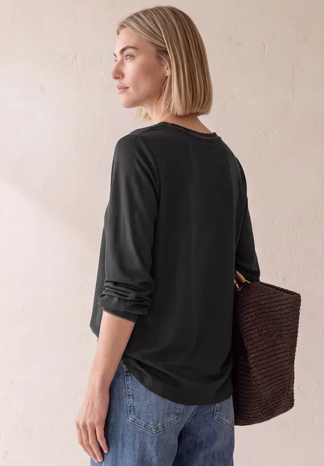 2-in-1 Sirt aus Chiffon mit Boat Neck Black Frau in schwarzem 3/4-Oberteil mit hochgekrempelten Ärmeln trägt Korbtasche und blickt seitlich.