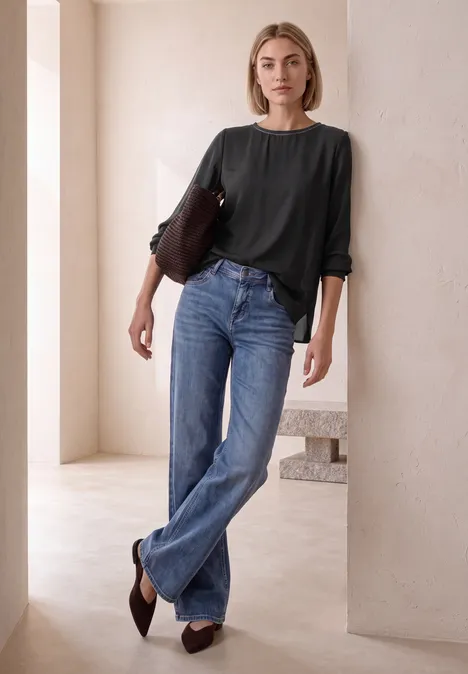 2-in-1 Sirt aus Chiffon mit Boat Neck Black Eine Frau in Jeans und schwarzem Oberteil lehnt locker an einer Säule in einem hellen, modernen Raum.