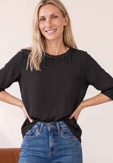 2-in-1 Shirt aus Chiffon mit Boat Neck black coffee Lächelnde Frau mit schwarzem T-Shirt und Jeans, steht mit Händen in der Hüfte vor neutralem Hintergrund.