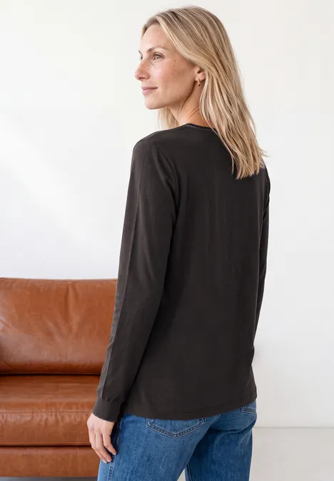 2-in-1 Shirt aus Chiffon mit Boat Neck black coffee Blonde Frau trägt schwarzes Langarmshirt und blaue Jeans, steht seitlich vor einem braunen Sofa, Blick nach links.