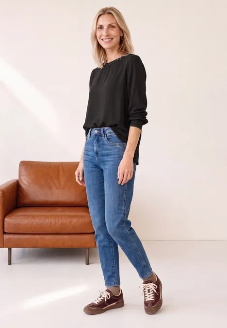 2-in-1 Shirt aus Chiffon mit Boat Neck black coffee Eine lächelnde Frau in schwarzer Bluse und blauen Jeans steht vor einem braunen Sofa.