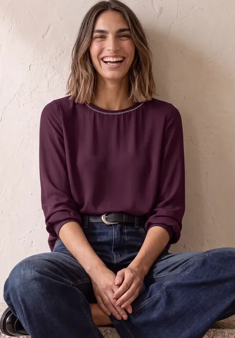 2-in-1 Sirt aus Chiffon mit Boat Neck berry maroon Eine lächelnde Frau in bordeauxrotem Pullover und Jeans sitzt entspannt mit verschränkten Beinen.