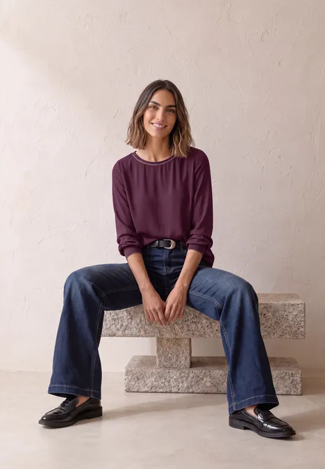 2-in-1 Sirt aus Chiffon mit Boat Neck berry maroon Eine Frau mit lila Oberteil und weitem Jeans sitzt entspannt auf einer steinernen Bank vor hellem Hintergrund.