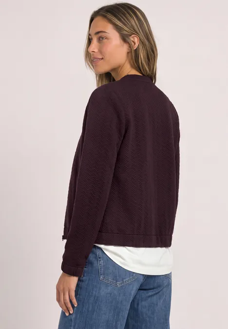 Leichte Sweatjacke mit Struktur und Zipper berry plum Junge Frau im Profil blickt über die Schulter, trägt dunklen Cardigan, weißen Pullover und blaue Jeans.
