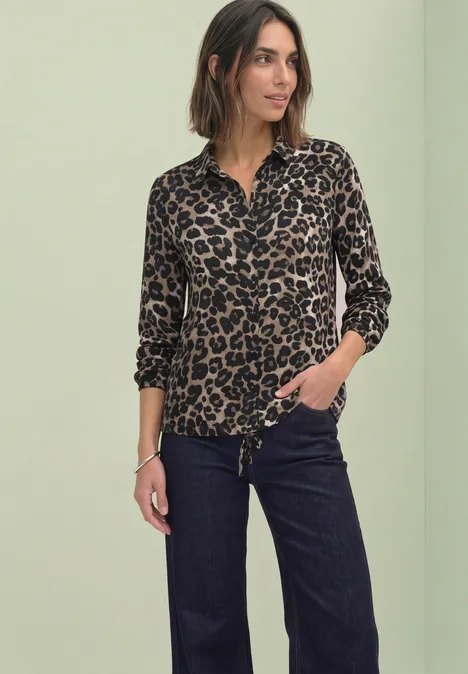 Frau in Leopardenmuster-Bluse und dunklen Jeans vor grünem Hintergrund.