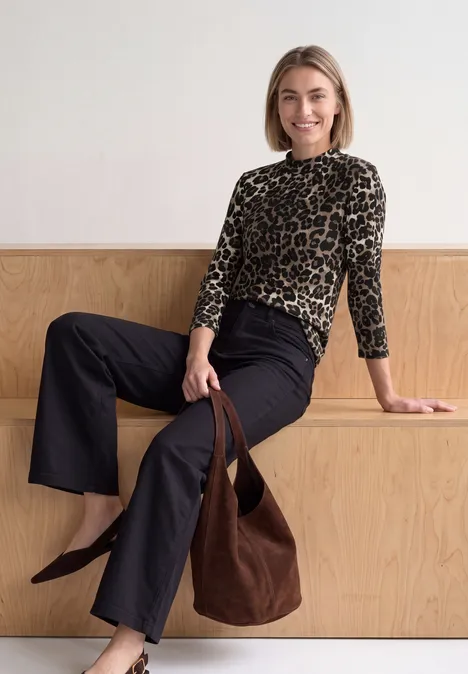Une femme sourit assise sur une structure en bois, en pull léopard, pantalon noir et sac marron.