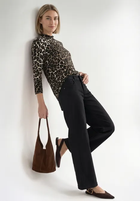 Femme appuyée contre un mur blanc, porte un top léopard, pantalon noir, sac et mocassins marron.