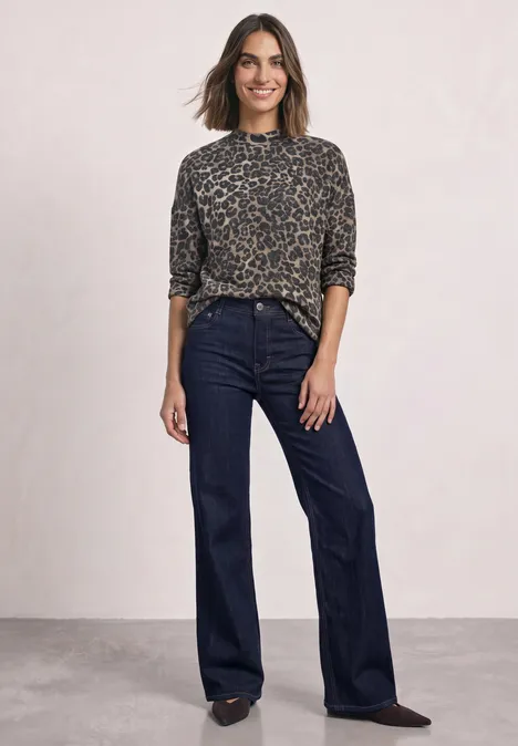 Langarm Shirt mit Turtle neck und Leo-Muster Black Frau in Leopardenbluse und dunklen Schlagjeans lächelt vor neutralem Hintergrund.