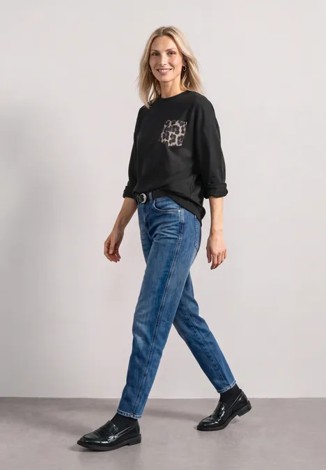 Een vrouw in zwarte trui, blauwe jeans en zwarte schoenen loopt in een neutrale ruimte.