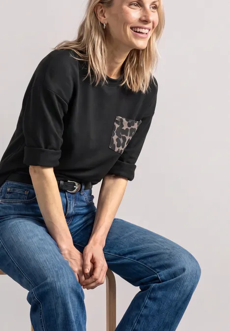 Glimlachende blonde vrouw in zwart shirt met luipaardzak en blauwe jeans, ontspannen zittend op een kruk.