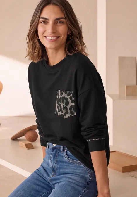 Langarm Shirt mit Leo-Details Black Frau in schwarzem Sweatshirt mit Leoprint-Tasche und blauer Jeans lächelt vor neutralem Hintergrund.