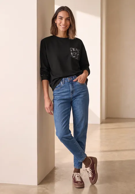 Langarm Shirt mit Leo-Details Black Eine Frau lehnt entspannt an einer Säule, trägt schwarzes Shirt mit Brusttasche, blaue Jeans und braune Schuhe.