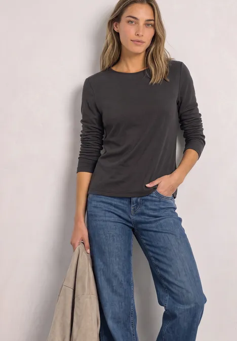 Shirt met lange mouwen, ronde hals en satijndetail black coffee Vrouw in zwart shirt met lange mouwen, blauwe jeans, houdt een lichte jas vast voor een lichte achtergrond.