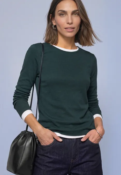 Shirt met lange mouwen, ronde hals en satijndetail stormy green Vrouw in donkergroene trui over wit shirt en spijkerbroek, zwarte schoudertas, kijkt in de camera.