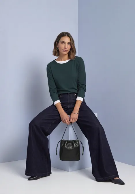 Shirt met lange mouwen, ronde hals en satijndetail stormy green Vrouw in donkergroene trui, wijde broek en zwarte schoenen zit, houdt een zwarte handtas vast.