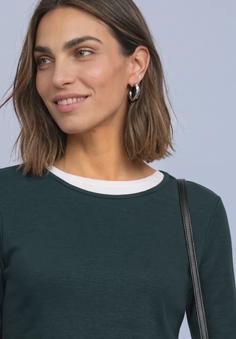 Shirt met lange mouwen, ronde hals en satijndetail stormy green Een glimlachende vrouw met halflang bruin haar, groen shirt met witte kraag en een tas over de schouder.