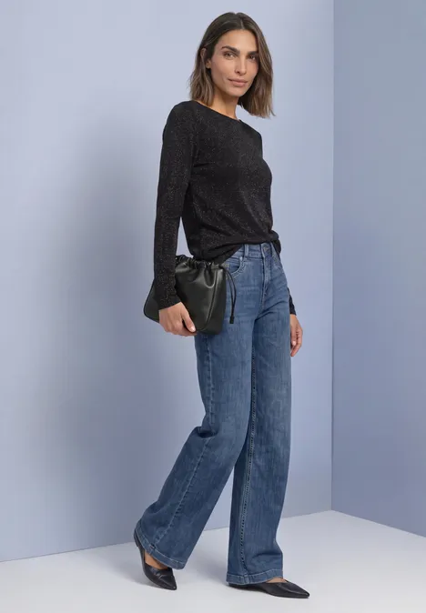 Shirt met lange mouwen, boothals en glitters Black Frau in schwarzem Oberteil, weitem Jeans, spitzen Schuhen, hält eine schwarze Clutch vor hellgrauer Wand.