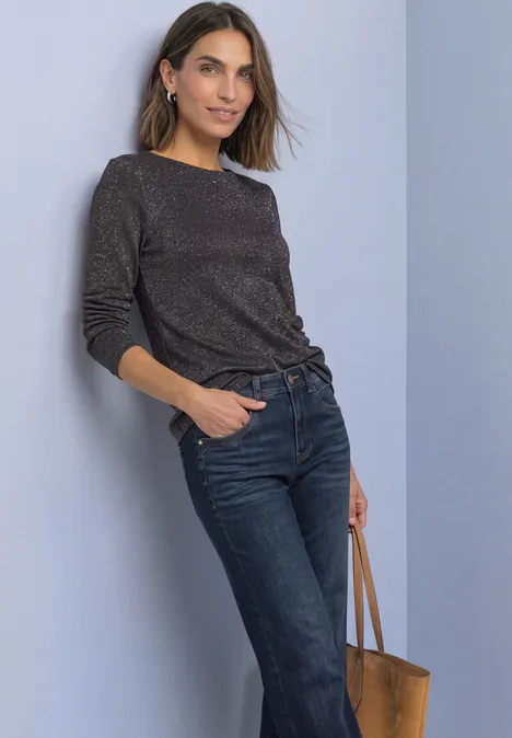 Langarm Shirt mit Boat Neck und Glitzer black coffee Frau lehnt an Wand, trägt schwarzen Pullover, blaue Jeans und hält eine braune Ledertasche.