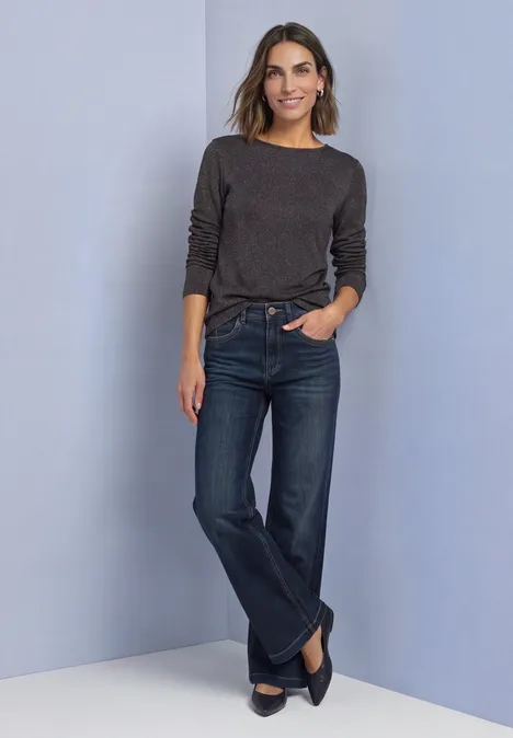 Langarm Shirt mit Boat Neck und Glitzer black coffee Frau lehnt an hellblauer Wand, trägt dunkelgraues Longsleeve, gerade geschnittene Jeans und schwarze Ballerinas.
