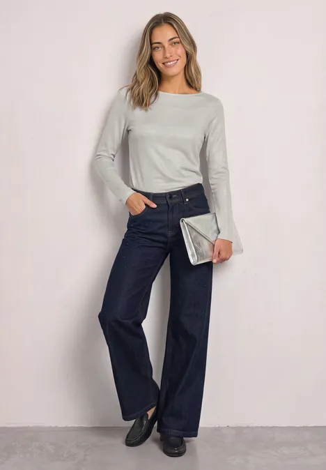 T-shirt à manches longues, col bateau et paillettes pure beige Frau mit grauem Langarmshirt, weiten Jeans und silberner Clutch, lächelnd vor hellem Hintergrund.