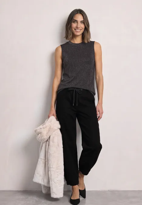 Top met col en glitters black coffee Vrouw in donkergrijze mouwloze top, zwarte joggingbroek en ballerina’s, houdt losjes een lichte mantel in haar hand