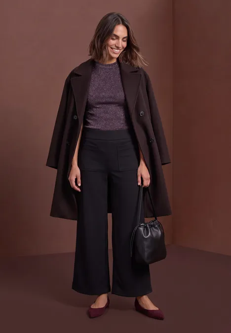 Top met col en glitters berry plum Vrouw met bruine mantel over de schouders, paarse top, wijde zwarte broek, platte schoenen en tas.