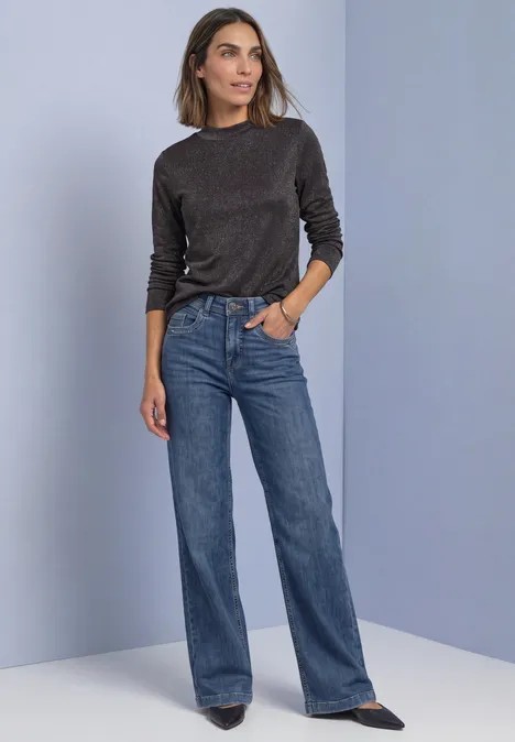 Shirt met lange mouwen, col en glitters black coffee Vrouw in zwarte longsleeve, wijde blauwe jeans en zwarte schoenen voor paarse achtergrond.