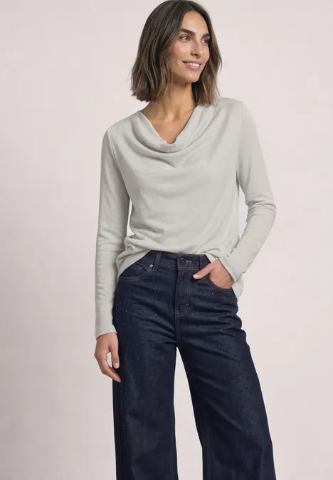 T-shirt à manches longues avec encolure en cascade pure beige Frau trägt graues Langarmshirt mit Wasserfallausschnitt und dunkle Jeans, steht lächelnd vor hellem Hintergrund.