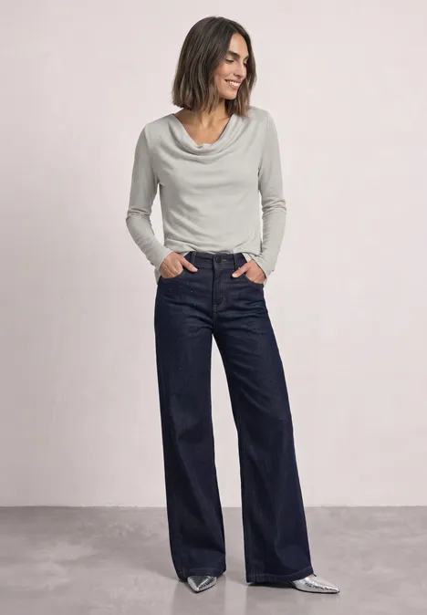 T-shirt à manches longues avec encolure en cascade pure beige Frau in weißem Wasserfallkragenoberteil und weiten dunkelblauen Jeans, Hände in den Taschen, silberne Schuhe.