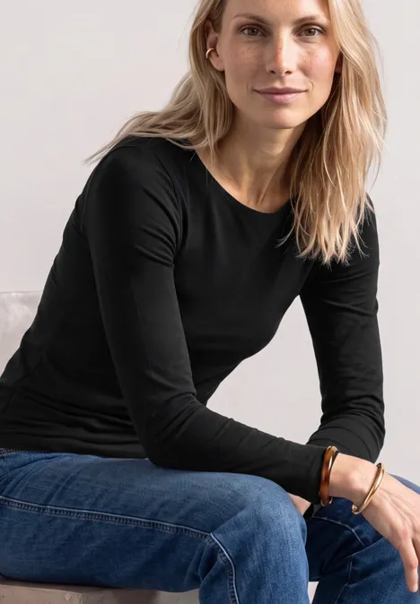 Basic shirt met boothals Black Eine Frau mit langen blonden Haaren in schwarzem Oberteil und blauer Jeans sitzt entspannt.