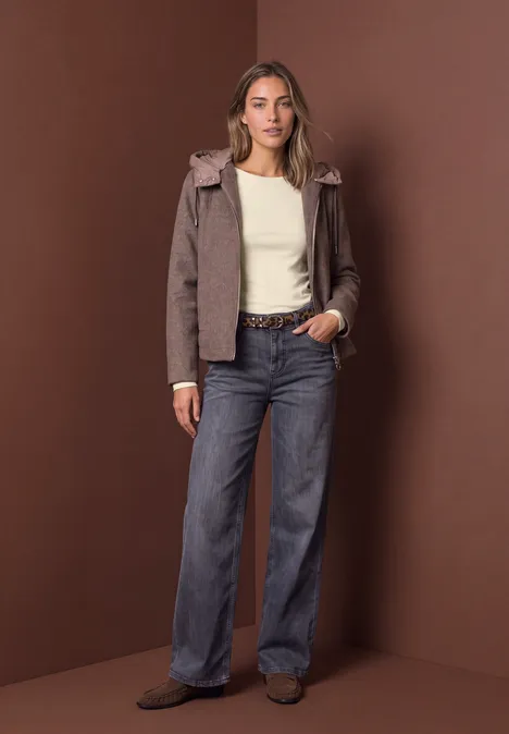 Femme portant une veste brune, un top blanc et un jean ample, posant devant un fond marron en studio.